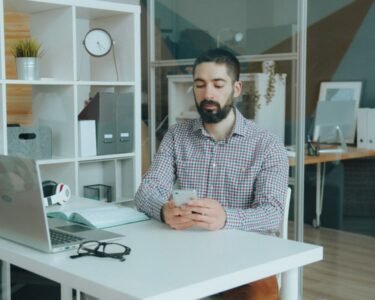 A man in a modern office setting using a smartphone at his desk, surrounded by office supplies.