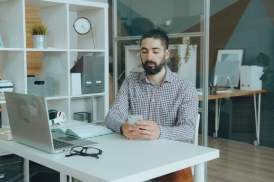 A man in a modern office setting using a smartphone at his desk, surrounded by office supplies.