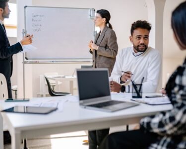 A diverse group of professionals engaged in a collaborative office meeting with laptops and a whiteboard.